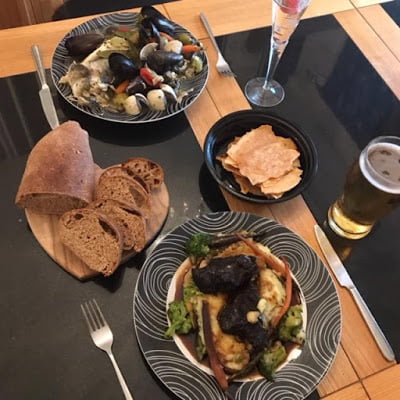 Two dishes prepared by Shardana Catering. One dish has braised slow cooked Ox cheek with smoked mashed potatoes and root vegetables, beef jus and baked seabass en papillote with seasonal veg. The other dish has mussels and herbs.